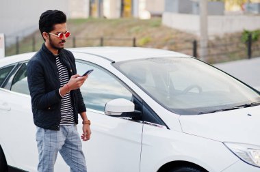 Güney Asyalı ya da Hintli erkek kırmızı gözlük takıyor. Yanında cep telefonu olan beyaz aracının yanında duruyor..