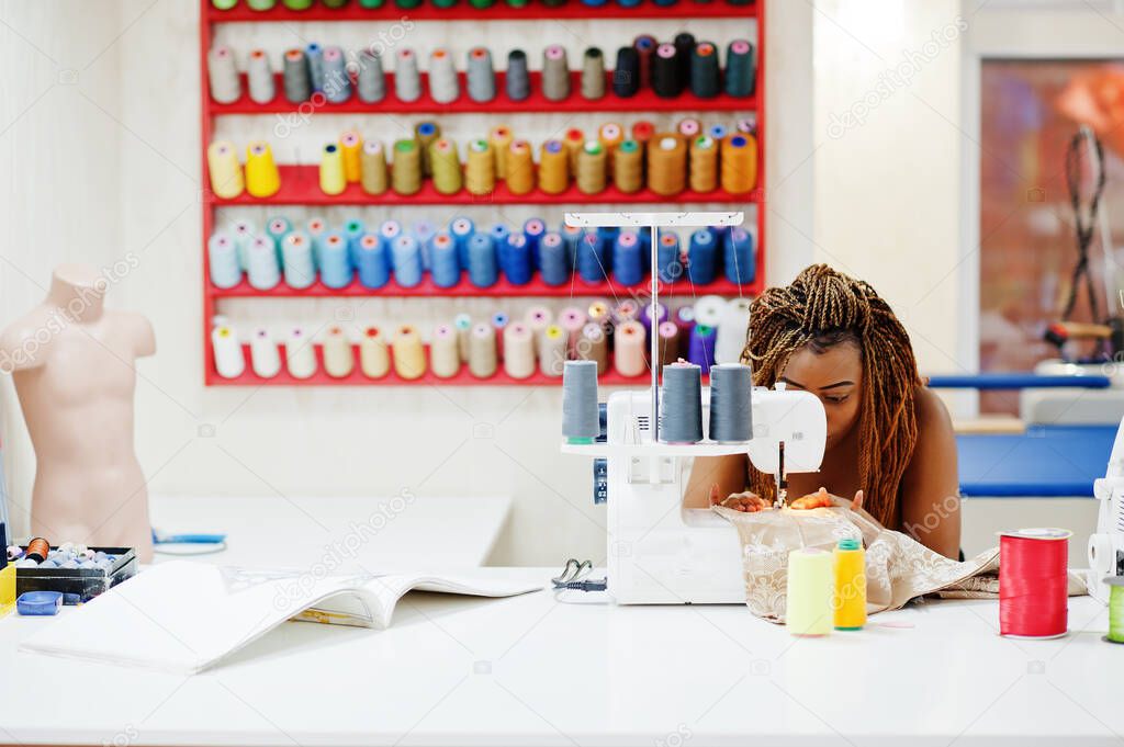 Young african dressmaker woman sews clothes on sewing machine at tailor office. Black seamstress women.