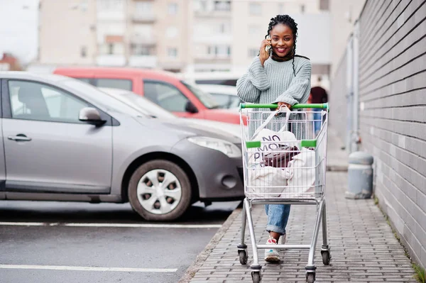 African woman with shopping cart trolley posed outdoor market near car parking and speaking on phone.