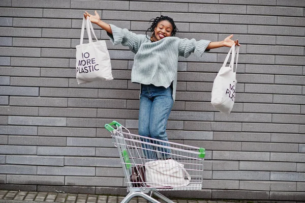 No more plastic. African woman with shopping cart trolley and eco bags jump outdoor market.