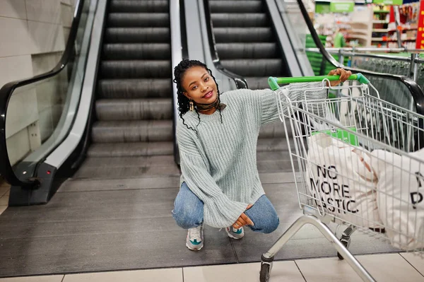 No more plastic. African woman with shopping cart trolley posed at supermarket against escalator stairs.
