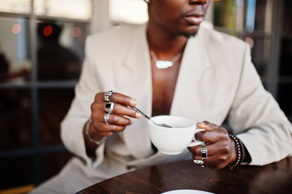 ベージュのスクールスーツを着たスタイリッシュなアフロマンがコーヒーを飲みながらカフェに座っています ファッショナブルな若いですアフリカの男性でカジュアルジャケットオン裸胴 — ストック写真
