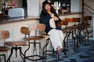 Çekici Afrikalı Amerikalı kadın manken siyah ceketli, şapkalı ve bel çantalı boş zamanlarında kafede dinleniyor..