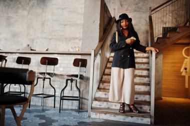 Çekici Afrikalı Amerikalı kadın model siyah ceketli, şapkalı ve bel çantalı boş zamanlarında kafede cep telefonuyla dinleniyor..