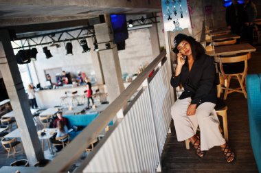 Çekici Afrikalı Amerikalı siyah ceketli kadın model boş zamanlarında kafede cep telefonuyla dinleniyor..