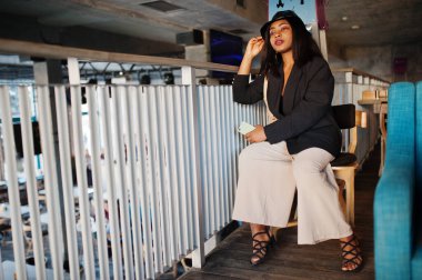 Çekici Afrikalı Amerikalı siyah ceketli kadın model boş zamanlarında kafede cep telefonuyla dinleniyor..