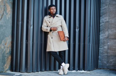 Siyah şapkalı ve bej ceketli, elinde klasörle dışarıda poz veren yakışıklı Afrikalı Amerikalı adam..