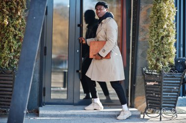 Siyah şapkalı ve bej ceketli, elinde klasörle dışarıda poz veren yakışıklı Afrikalı Amerikalı adam..