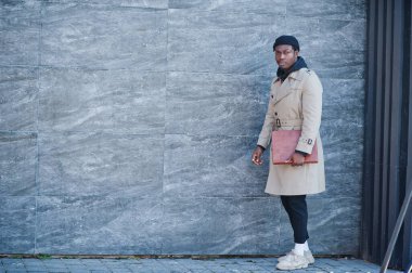 Siyah şapkalı ve bej ceketli, elinde klasörle dışarıda poz veren yakışıklı Afrikalı Amerikalı adam..