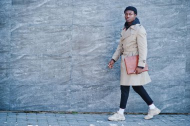 Siyah şapkalı ve bej ceketli, elinde klasörle dışarıda poz veren yakışıklı Afrikalı Amerikalı adam..