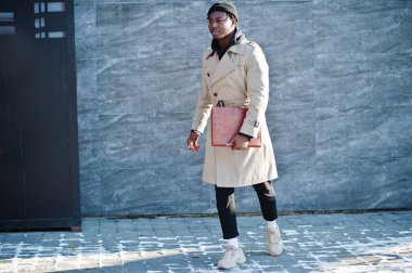 Siyah şapkalı ve bej ceketli, elinde klasörle dışarıda poz veren yakışıklı Afrikalı Amerikalı adam..