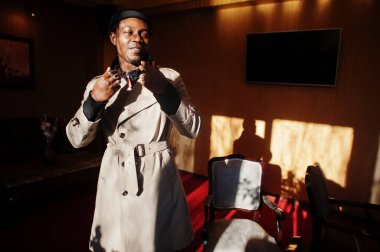 Siyah şapkalı ve bej ceketli, güneş ışığı gölgeli, içeride poz veren yakışıklı Afrikalı Amerikalı adam..