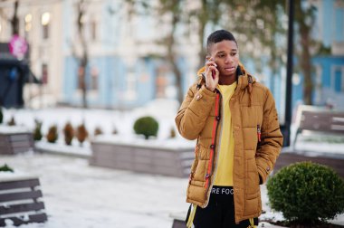 Afrikalı adam kış mevsiminde turuncu ceket giyer soğuk hava açık havada cep telefonuyla poz verir.
