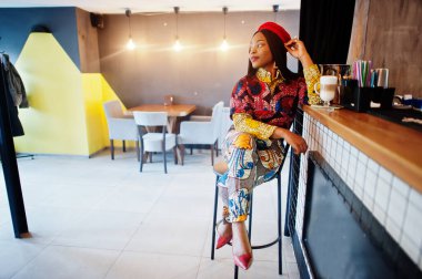 Coşkulu Afro-Amerikalı kadın moda renkli kıyafetli, kırmızı bereli, rahat bir kafede takılıyor, tezgahta oturuyor..