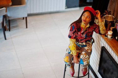 Coşkulu Afro-Amerikalı kadın moda renkli kıyafetli, kırmızı bereli, rahat bir kafede takılıyor, tezgahta oturuyor..