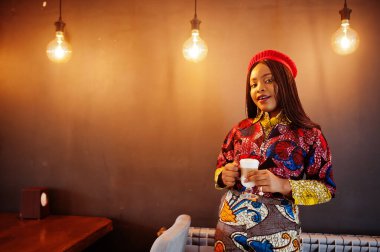 Coşkulu Afro-Amerikan kadın moda renkli kıyafetli, kırmızı bereli, elinde bir fincan sıcak latte ile rahat bir kafede takılıyor..