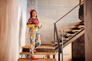 Coşkulu Afro-Amerikalı kadın moda renkli kıyafetli, rahat kafede kırmızı bereli, elinde bir bardak sıcak içecekle merdivenlerde duruyor..