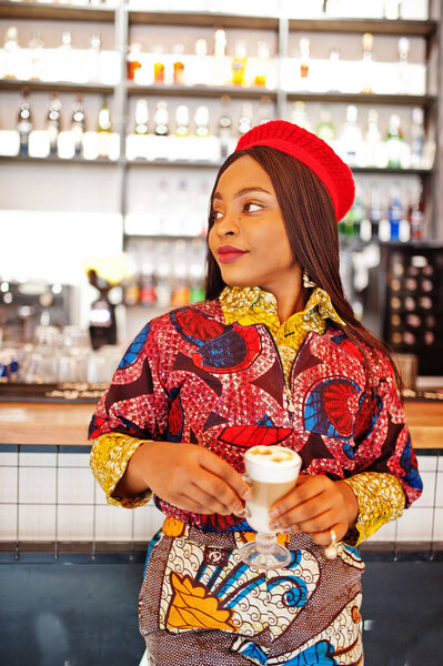 Enthusiastic african american woman in trendy coloured outfit with red beret chilling in cozy cafe, standing near bar counter.