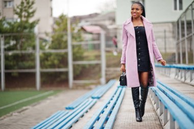 Stadyum tribünlerinde şık, güzel, afro-amerikan bir kadın. Moda kıyafeti giymiş, el çantalı..
