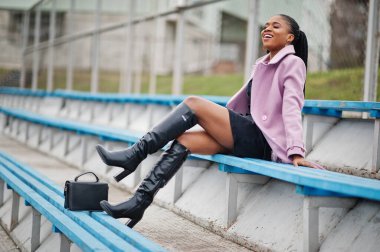 Stadyum tribünlerinde şık, güzel, afro-amerikan bir kadın. Moda kıyafeti giymiş, el çantalı..