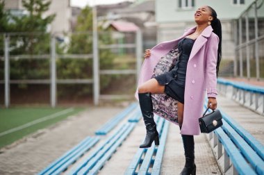 Stadyum tribünlerinde şık, güzel, afro-amerikan bir kadın. Moda kıyafeti giymiş, el çantalı..