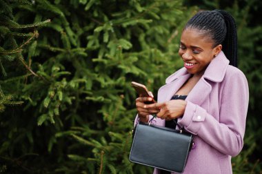 Genç, şık, Afro-Amerikalı bir kadın çam ağaçlarının yanında, moda kıyafeti giymiş, el çantası ve cep telefonu olan..