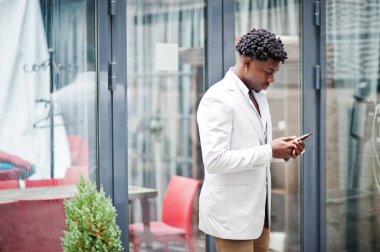 Düşünceli, genç, yakışıklı, Afro-Amerikalı, resmi giysili bir beyefendi. Beyaz ceketli siyahi model cep telefonuna bakıyor..