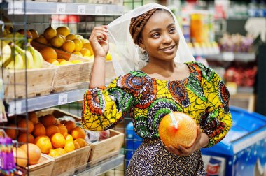 Geleneksel giysiler ve duvak giyen mutlu Afrikalı kadın markette ürün arıyor, süpermarketten alışveriş yapıyor. Marketten meyve alan Afro siyah kadın kostümcü.