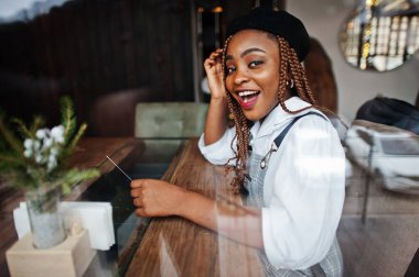 Önlüklü ve bereli Afro-Amerikalı kadın cam pencerenin önünde oturuyor..
