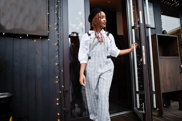 African american woman in overalls and beret walking out the door in outdoor terrace with christmas decorations.