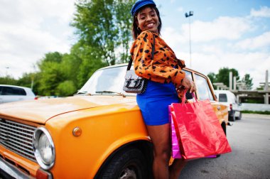 Güzel Afro-Amerikalı kadın, alışveriş torbalarıyla turuncu klasik eski arabanın yanında duruyor..