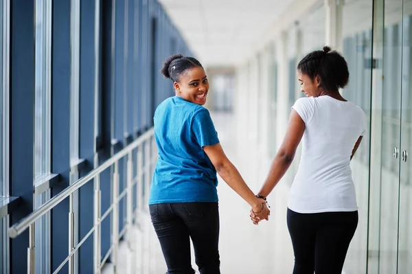 Back of two african woman friends in t-shirts walking halding hands indoor together.