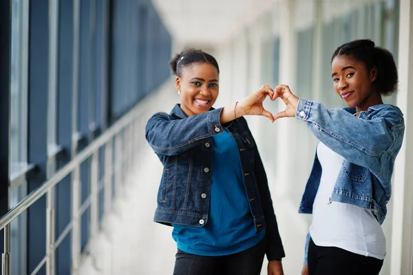 Two african woman friends in jeans jacket showing heart by fingers indoor together.