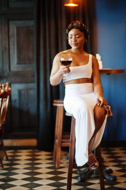 Afro-Amerikalı kadın, retro saç stili, beyaz elbiseli, restoranda bir bardak şarapla..