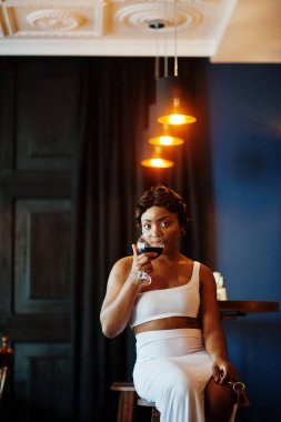 Afro-Amerikalı kadın, retro saç stili, beyaz elbiseli, restoranda bir bardak şarapla..