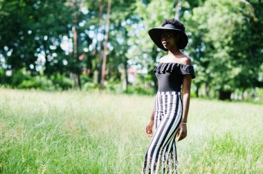 20 'li yaşlarda Afro-Amerikan güzel bir kadının portresi. Siyah beyaz pantolon ve yeşil çimlere poz veren yaz şapkası..