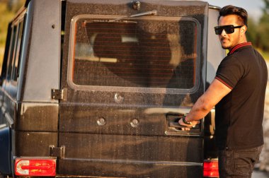 Asyalı adam siyah giyinmiş, SUV 'un yanında poz vermiş, arka kapı açık..