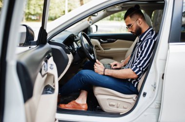 Başarılı bir Arap erkeği çizgili gömlek ve güneş gözlüğü takıyor ve elinde dizüstü bilgisayarla beyaz SUV arabasının direksiyonunda poz veriyor..