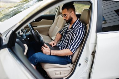 Başarılı bir Arap erkeği çizgili gömlek ve güneş gözlüğü takıyor ve elinde dizüstü bilgisayarla beyaz SUV arabasının direksiyonunda poz veriyor..