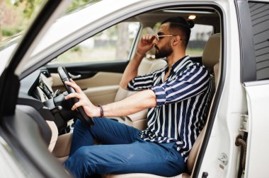 Başarılı Arap adam çizgili gömlek ve güneş gözlüğü takıyor beyaz SUV arabasının direksiyonunda poz veriyor. Tarz sahibi Arap erkekleri nakliye işinde.