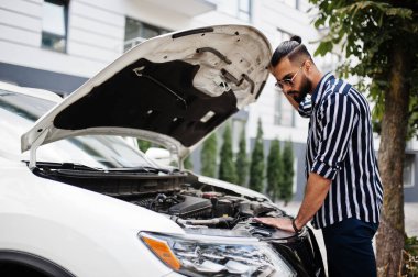 Başarılı Arap adam çizgili gömlek ve güneş gözlüğü takıyor beyaz SUV 'unun yanında poz veriyor, motoru açık kaputla kontrol ediyor..