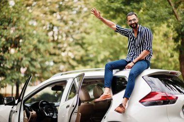 Başarılı bir Arap erkeği beyaz SUV arabasının çatısında oturan çizgili gömlek ve güneş gözlüğü takıyor..