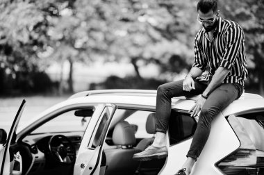 Başarılı bir Arap erkeği beyaz SUV arabasının çatısında oturan çizgili gömlek ve güneş gözlüğü takıyor..