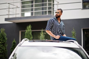 Başarılı bir Arap erkeği beyaz SUV arabasının çatısında oturan çizgili gömlek ve güneş gözlüğü takıyor..