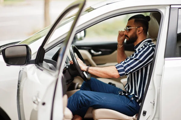 Başarılı Arap adam çizgili gömlek ve güneş gözlüğü takıyor beyaz SUV arabasının direksiyonunda poz veriyor. Tarz sahibi Arap erkekleri nakliye işinde.