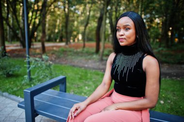 Şeftali pantolonlu ve siyah bluzlu modacı Afro-Amerikalı kadın dışarıda, bankta oturuyor..