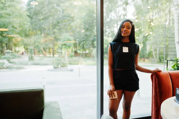 Fashionable feminist african american woman wear in black t-shirt and shorts, posed at restaurant.