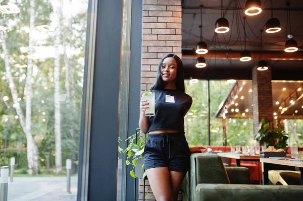 Fashionable feminist african american woman wear in black t-shirt and shorts, posed at restaurant with lemonade glass.