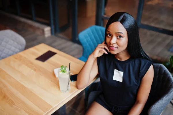 Fashionable feminist african american woman wear in black t-shirt and shorts, posed at restaurant with lemonade glass.