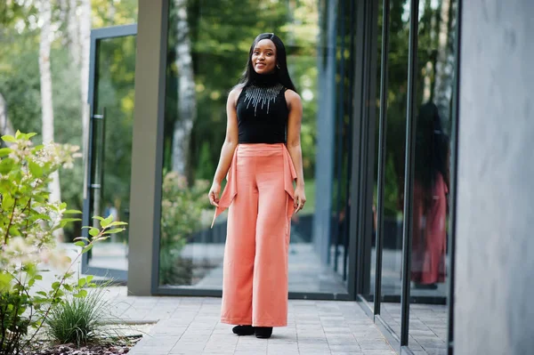 Fashionable african american woman in peach pants and black blouse pose outdoor.